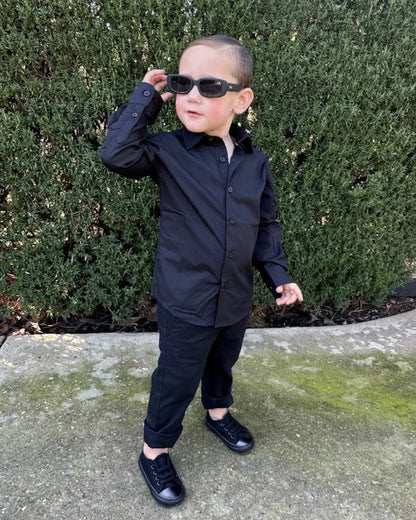Child wearing black shirt, chinos and sunglasses standing outdoors with greenery in the background